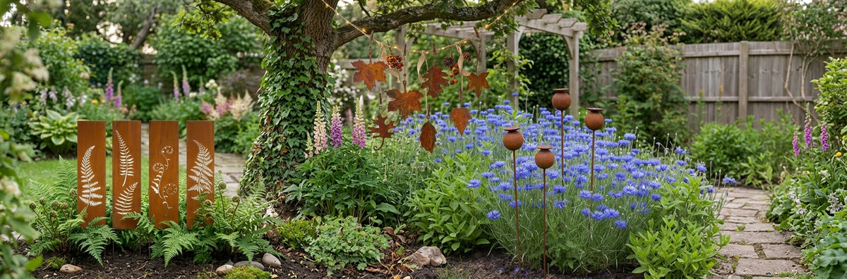 A landscaped garden bed with blue flowers, green foliage, a stone path, and several rust-colored metal garden stakes with decorative leaf and fern shapes, set under a tree near a wooden fence.