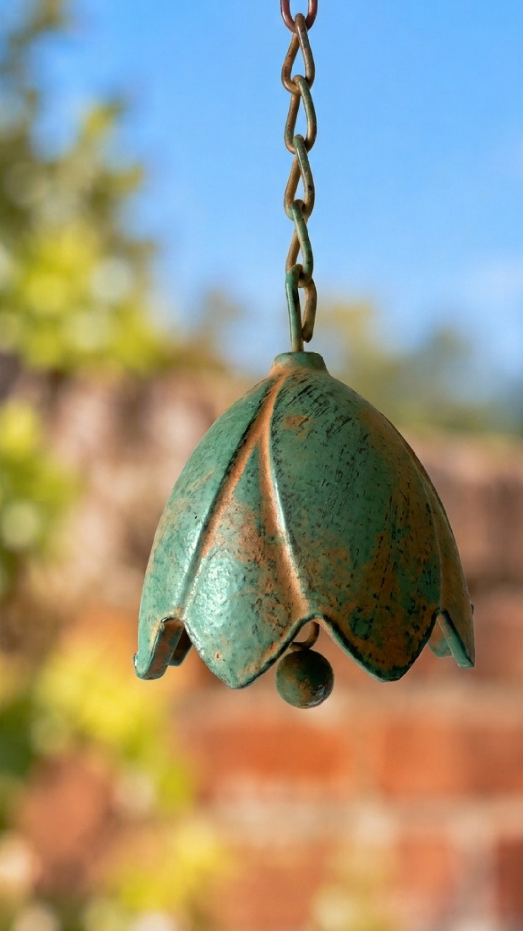 A hanging metal garden bell with a distressed green and brown patina finish, suspended by a chain against a blurred outdoor background of blue sky and foliage.